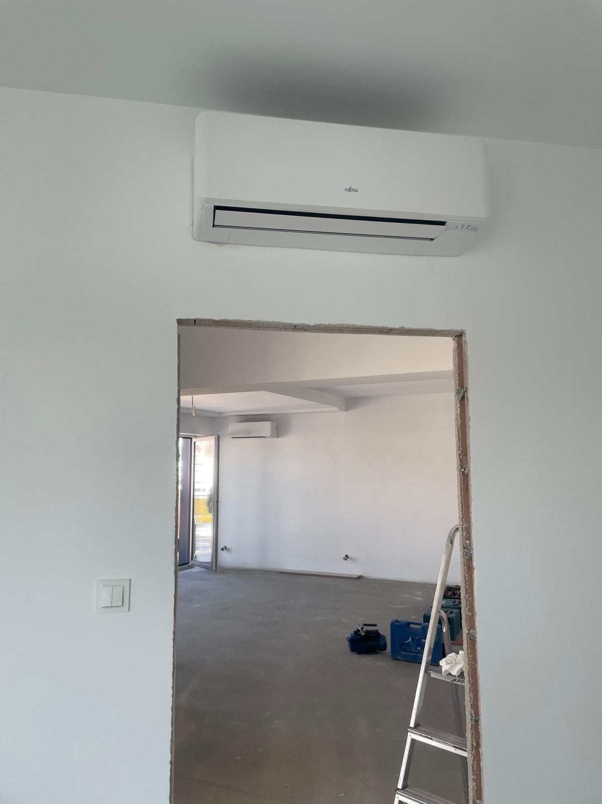 Biały klimatyzator Fujitsu zamontowany na ścianie nad surowym otworem drzwiowym w nowym, pustym pokoju z szarą podłogą; w tle widoczny drugi klimatyzator i drabina.