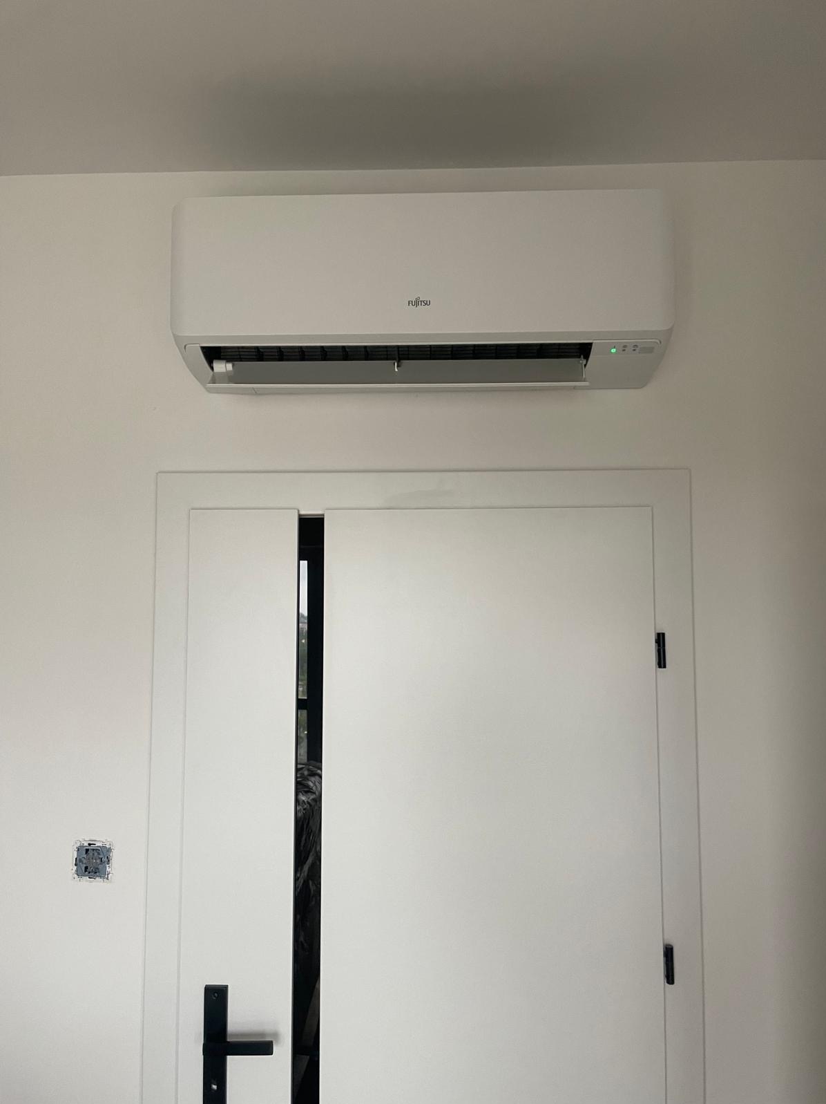 Biały klimatyzator Fujitsu zamontowany nad przesuwnymi drzwiami w minimalistycznym wnętrzu, widoczna puszka elektryczna bez osprzętu na ścianie.