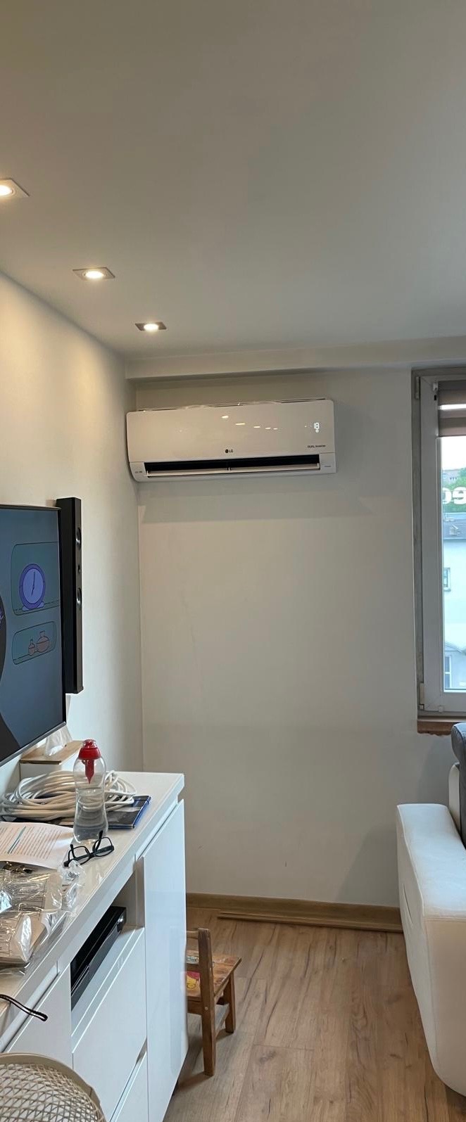 Biały klimatyzator LG zamontowany na ścianie w jasnym pokoju, widoczny fragment telewizora i okna z widokiem na budynek.