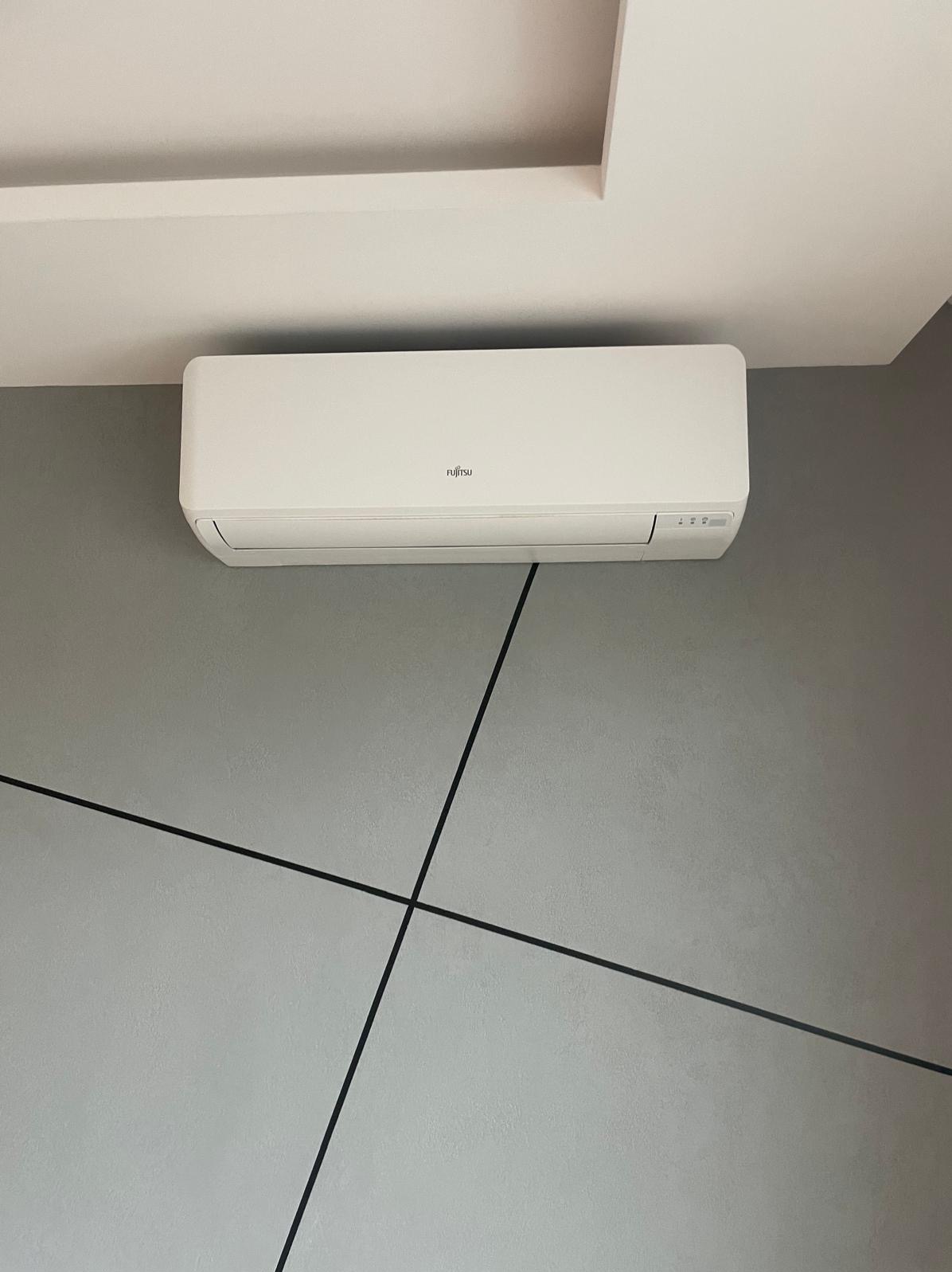 Biały klimatyzator marki Fujitsu zamontowany na szarej ścianie z dekoracyjnymi, czarnymi liniami, widok z dołu.