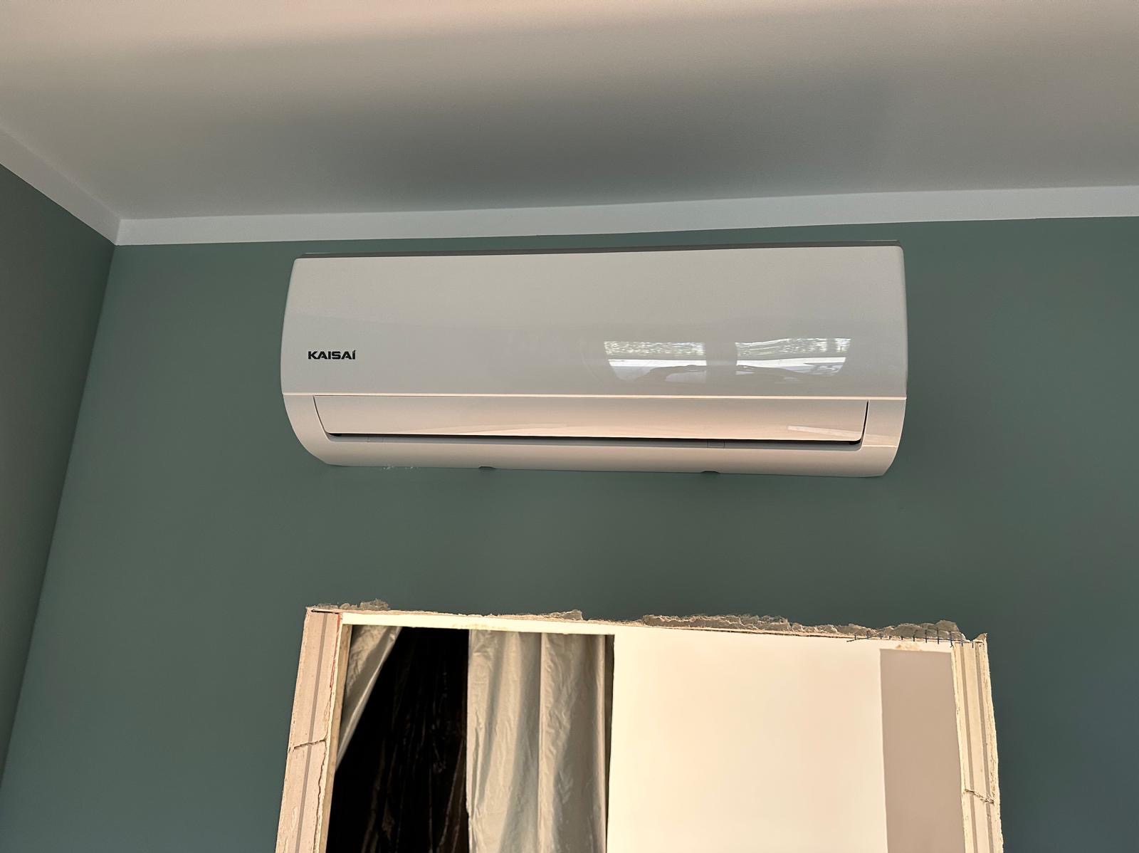 Biały klimatyzator Kaisai zamontowany na zielonej ścianie w pokoju, widoczny fragment otworu drzwiowego w trakcie remontu.