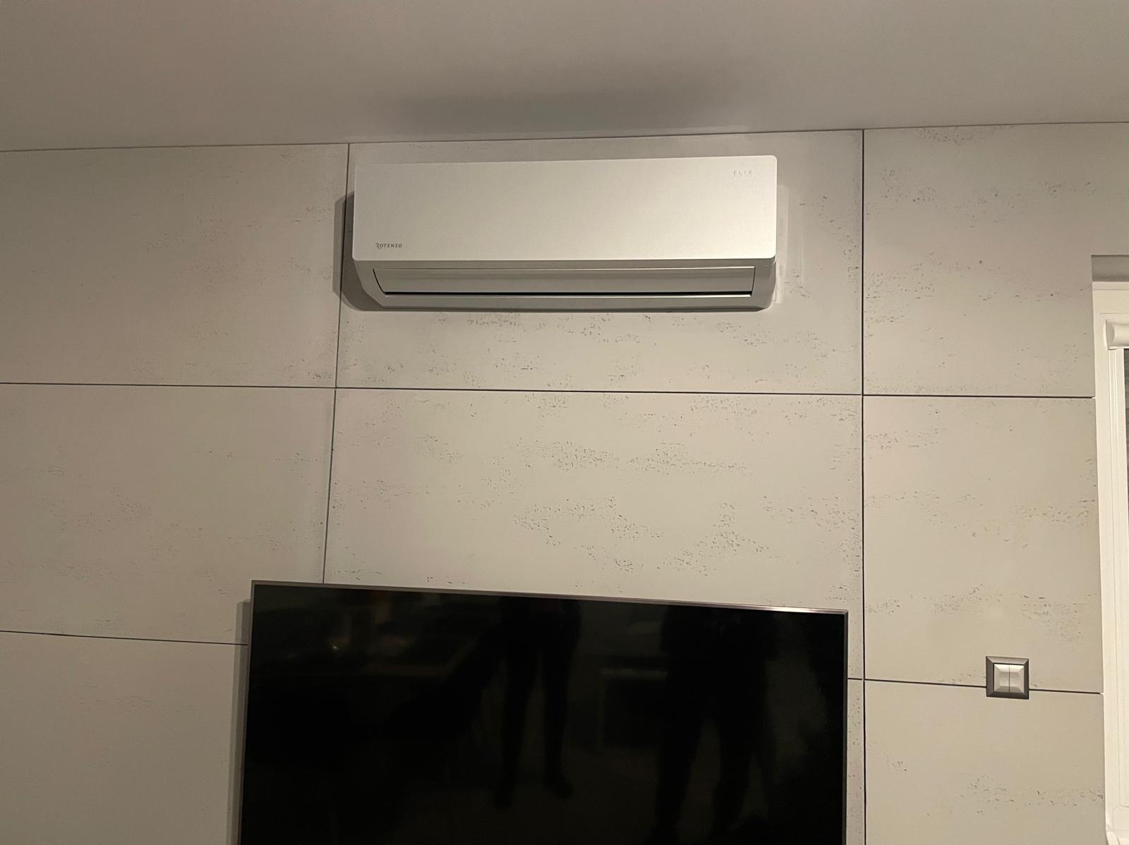 Zamontowany srebrny klimatyzator marki Rotenso na ścianie wyłożonej panelami imitującymi beton, pod nim widoczny telewizor.
