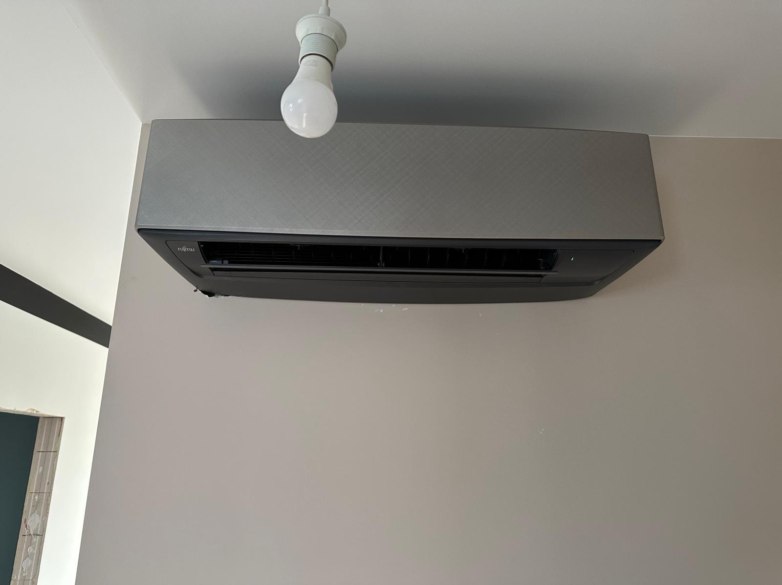 Szary klimatyzator firmy Fujitsu zamontowany na ścianie w jasnym pomieszczeniu, widoczna żarówka bez klosza.