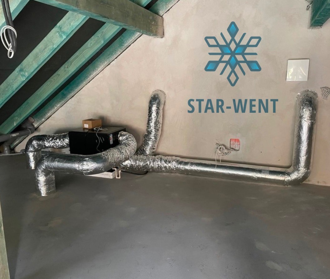 Zamontowany system rekuperacji z czarną centralą i srebrnymi, elastycznymi przewodami w pomieszczeniu na poddaszu z widoczną konstrukcją dachu i logo firmy STAR-WENT na ścianie.