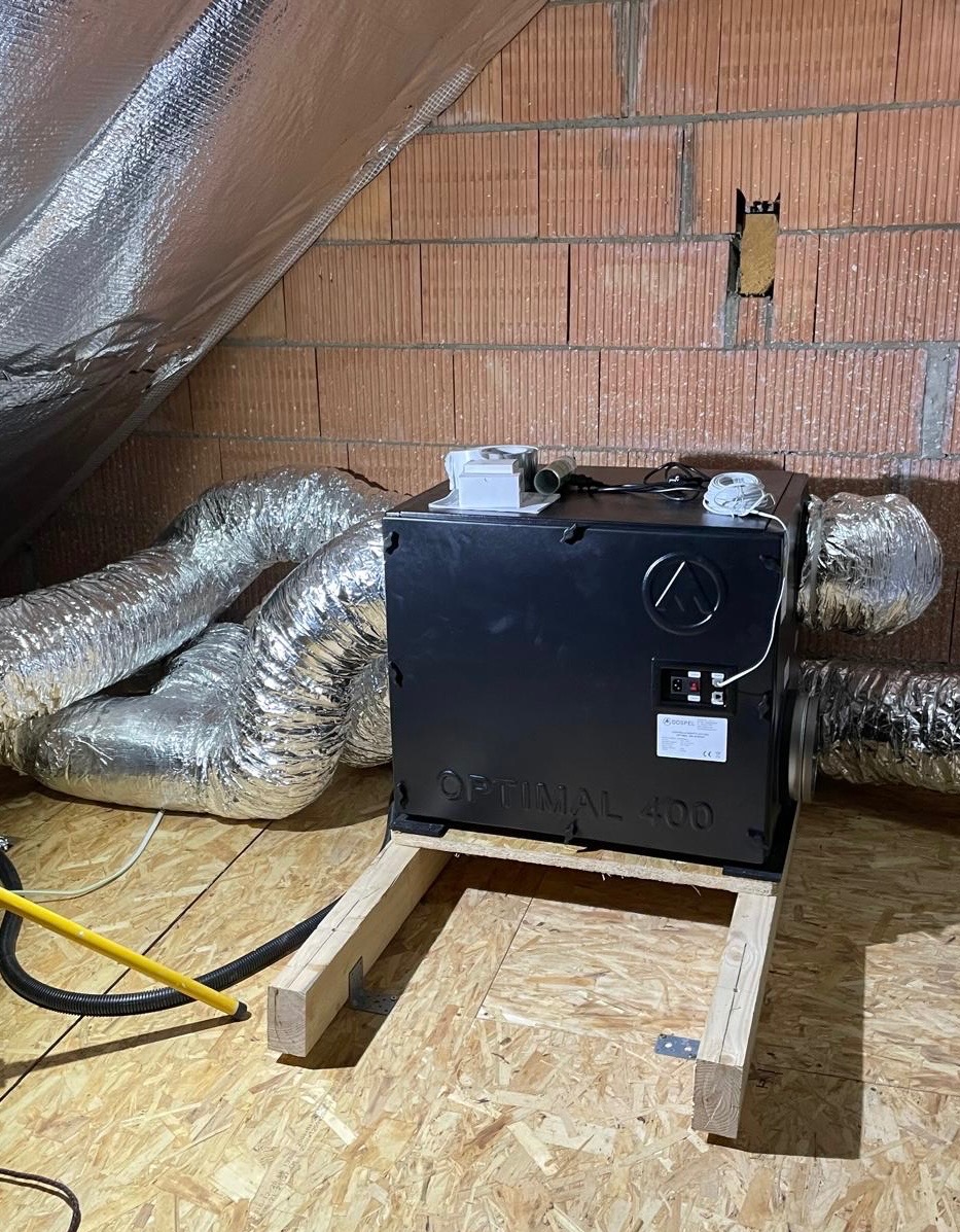 Czarna jednostka rekuperacyjna Optimal 400 zamontowana na drewnianej ramie w surowym pomieszczeniu na poddaszu z widocznymi przewodami wentylacyjnymi i izolacją folią aluminiową.