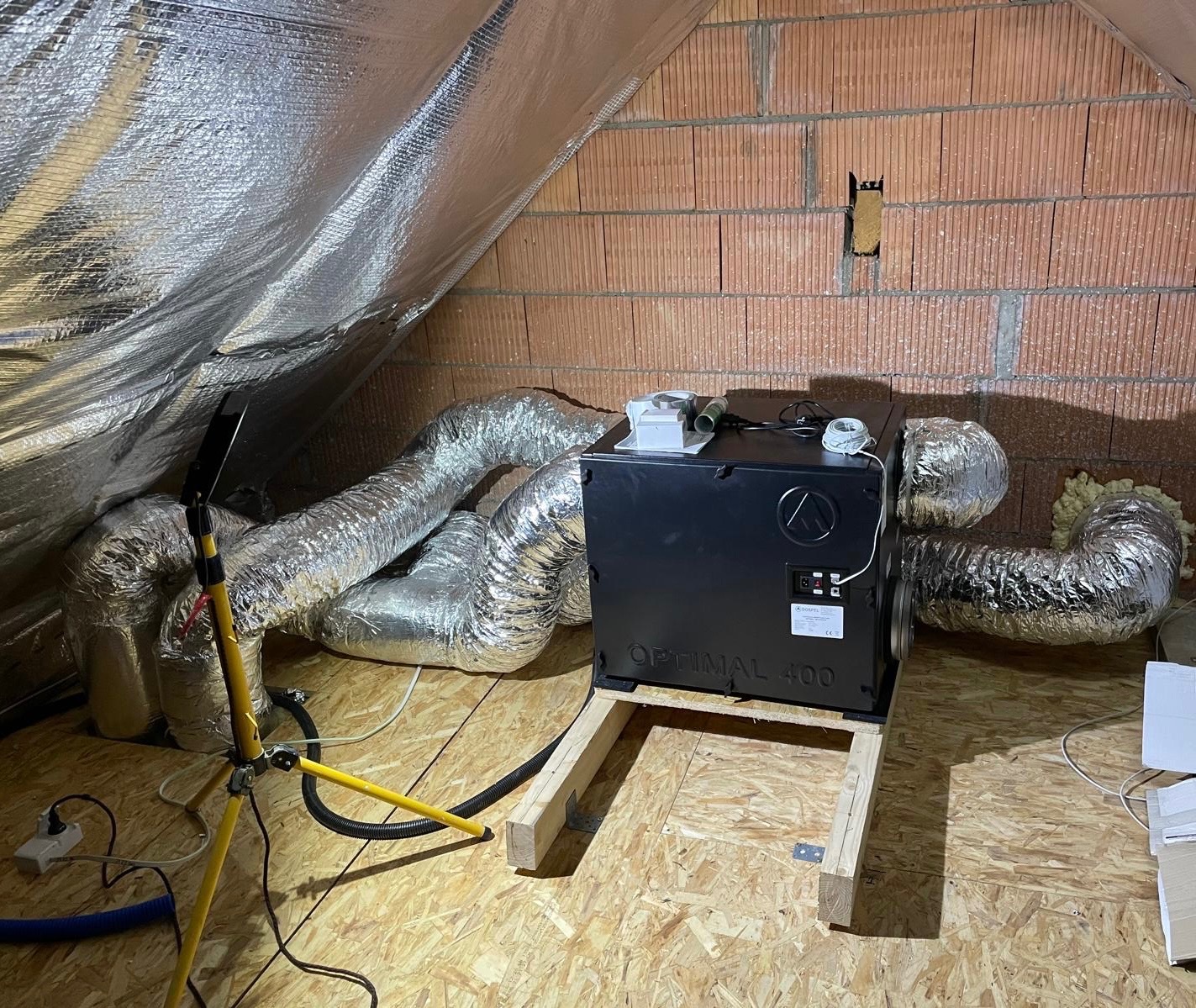 Czarna centrala wentylacyjna Optimal 400, zamontowana na drewnianej konstrukcji na poddaszu, z podłączonymi elastycznymi przewodami wentylacyjnymi, widoczna lampa robocza na trójnogu.