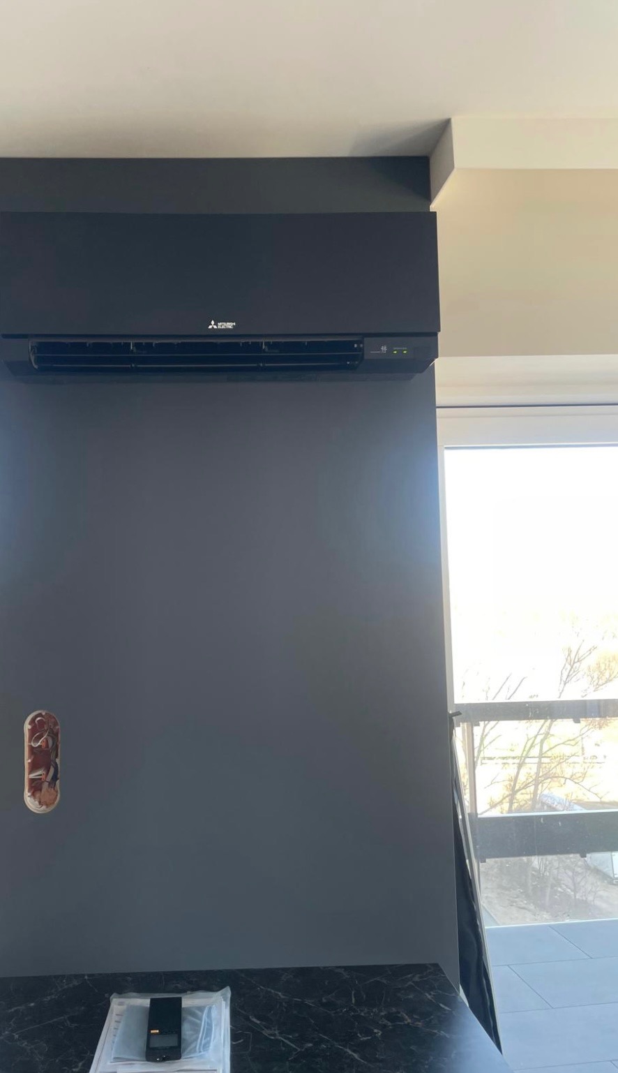 Czarny klimatyzator Mitsubishi zamontowany na szarej ścianie w nowoczesnym wnętrzu, widoczne przygotowane miejsce na gniazdko elektryczne i dokumentacja na blacie.