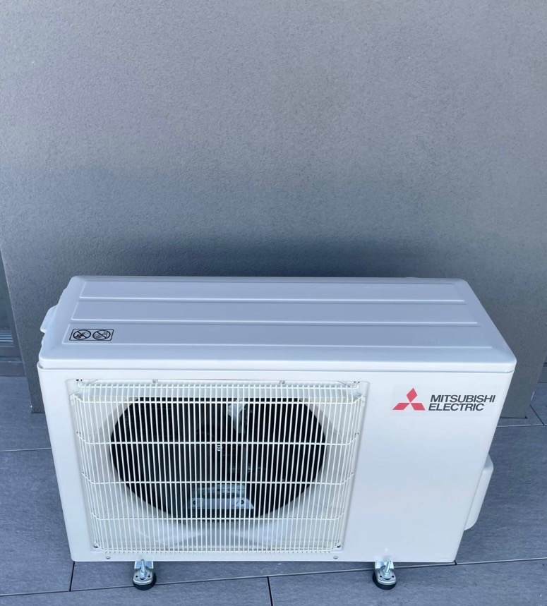 Biała, przenośna jednostka zewnętrzna klimatyzacji Mitsubishi Electric na kółkach, ustawiona na szarym podłożu, z widocznym logo producenta.