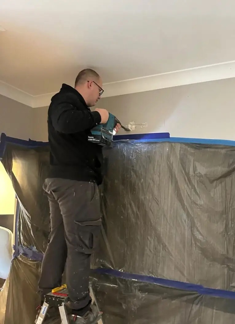 Pracownik na drabinie wierci otwór w ścianie, zabezpieczonej folią i taśmą malarską, używając wiertarki akumulatorowej Makita.