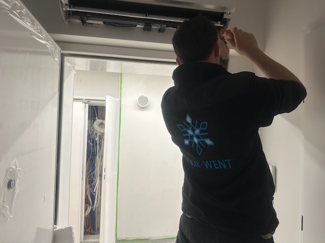 Instalator w bluzie z logo STAR-WENT montuje klimatyzację w pomieszczeniu z widocznymi kablami i otwartymi drzwiami.