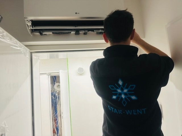 Instalator w firmowej bluzie STAR-WENT montuje białą klimatyzację LG na ścianie w pomieszczeniu z szafą elektryczną. Widoczne przewody w szafie.