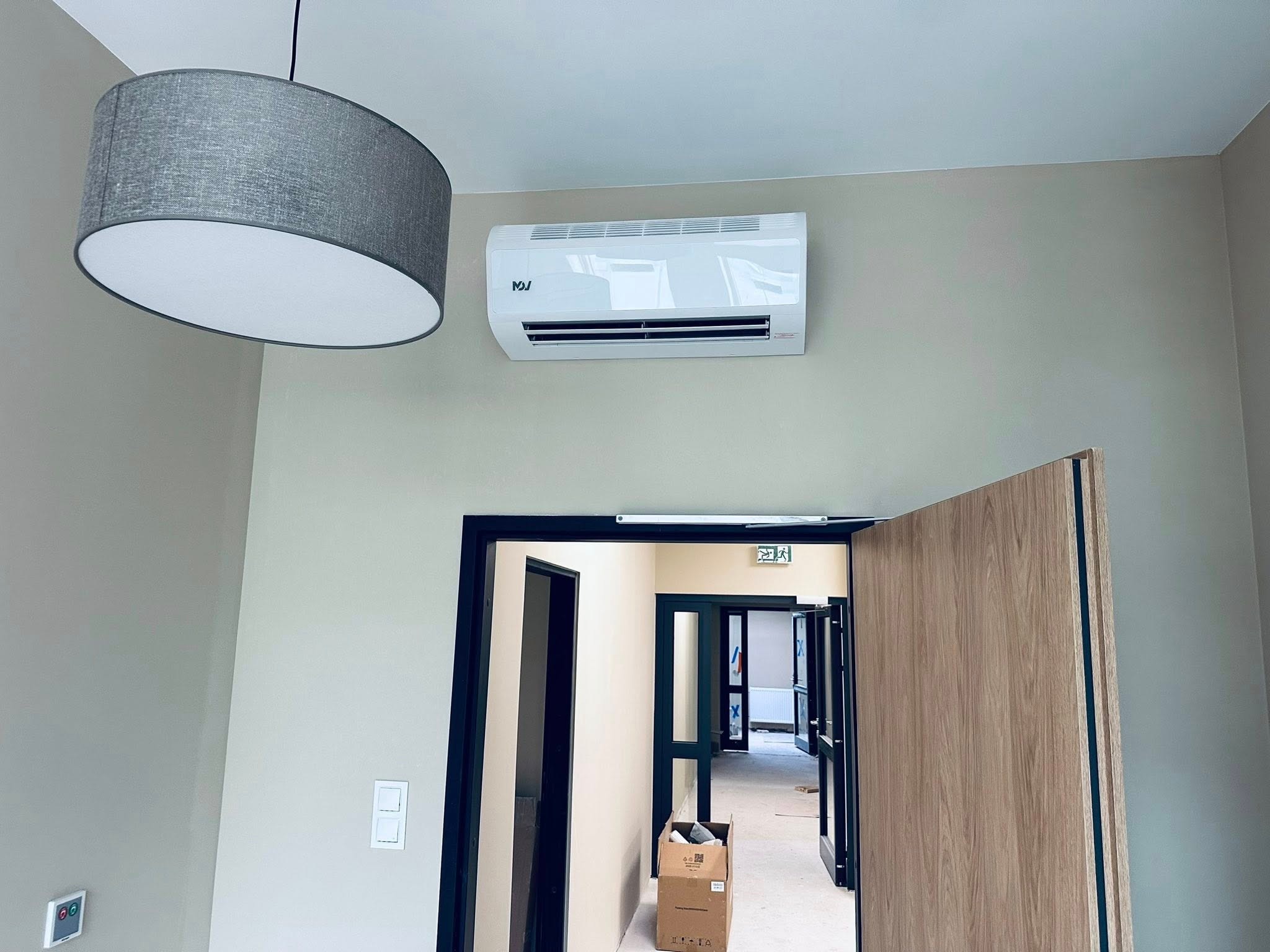 Biała klimatyzacja ścienna w jasnym wnętrzu, widoczna lampa sufitowa i otwarte drzwi do kolejnych pomieszczeń. Minimalistyczny design.