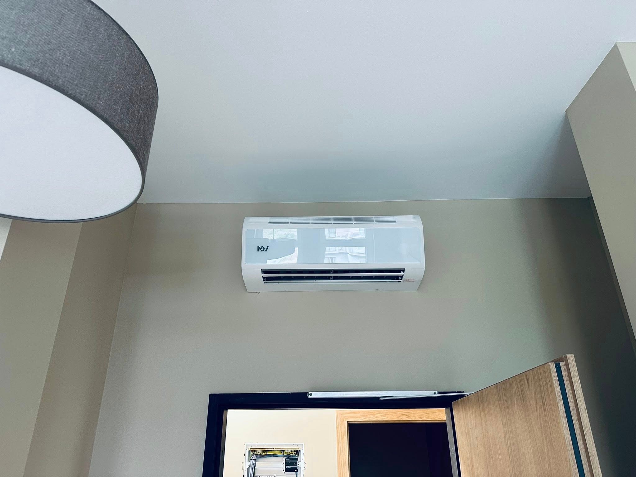 Biały klimatyzator ścienny nad otwartymi drzwiami w pokoju z beżowymi ścianami i szarą lampą sufitową. Widoczna skrzynka elektryczna w ścianie.