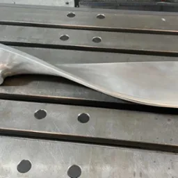 Metalowy, zakręcony element z otworem, leżący na stole frezarki. Widoczne ślady obróbki.