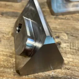 Precyzyjnie obrobiony, metalowy element o skomplikowanym kształcie, z widocznymi śladami frezowania na powierzchni, leżący na drewnianym blacie z wyraźną fakturą.