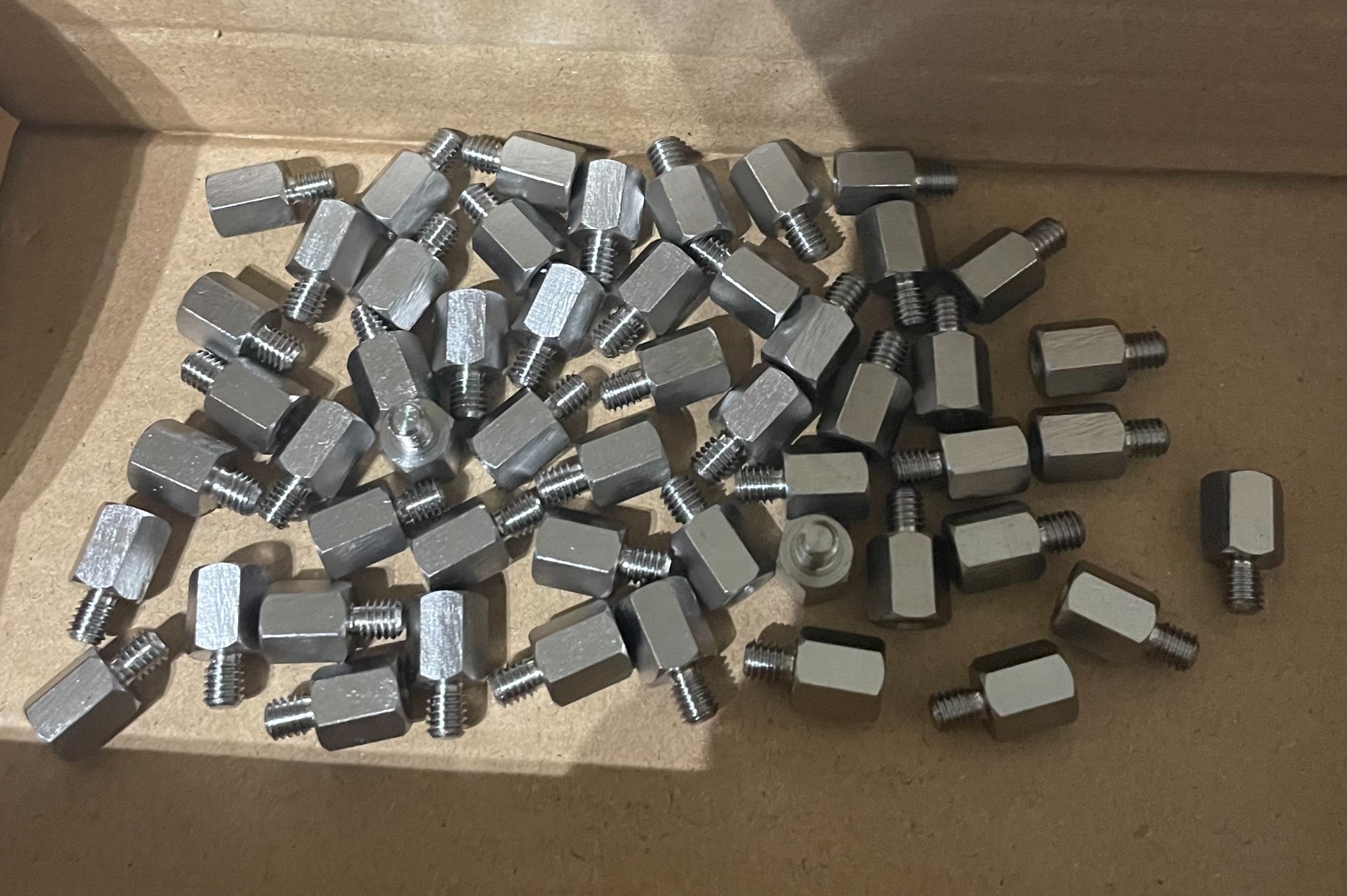 Partia srebrnych, metalowych dystansów gwintowanych w kartonowym pudełku, widoczne gwinty i sześciokątne korpusy.