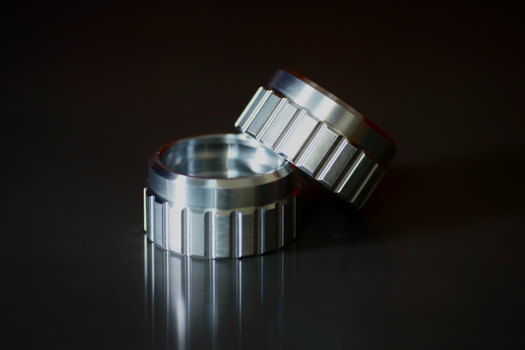 Dwa metalowe pierścienie z rowkowanymi krawędziami, ułożone jeden na drugim na błyszczącej, ciemnej powierzchni, tworzące odbicie.