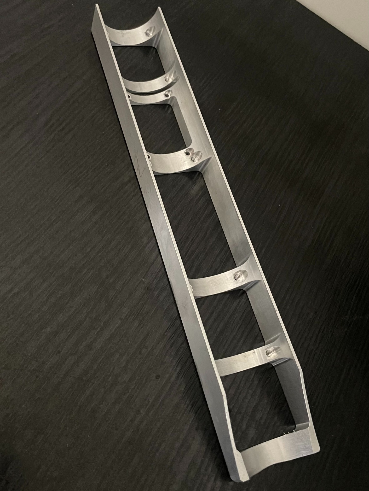 Aluminiowa rama z trzema półokrągłymi wycięciami, połączona poprzeczkami, leżąca na ciemnym, teksturowanym tle.