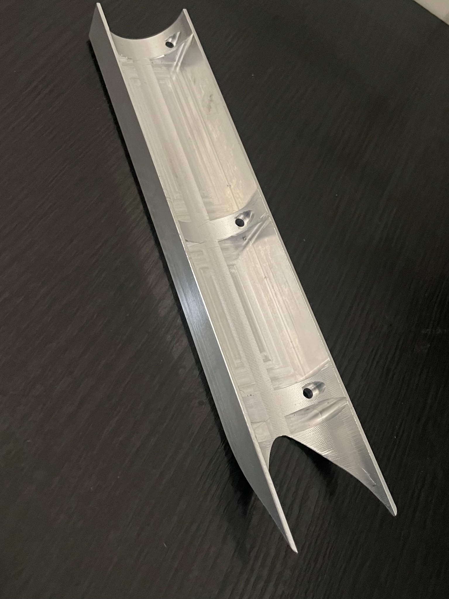 Aluminiowy profil o przekroju półksiężyca z otworami, po obróbce skrawaniem, leżący na ciemnym tle.