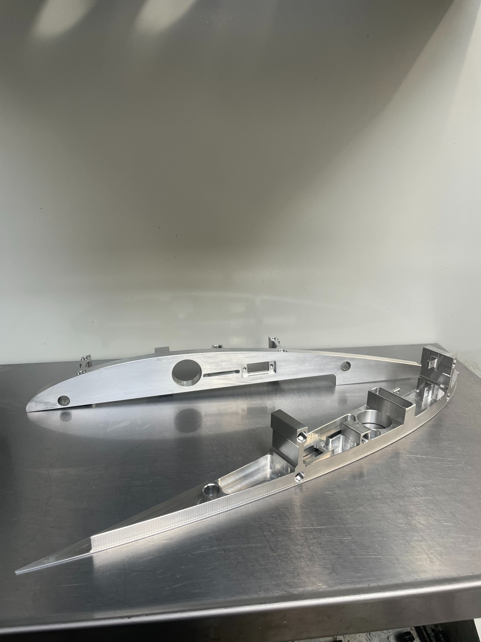 Dwie aluminiowe części obrobione z widocznymi otworami, wgłębieniami i detalami powierzchni, leżące na metalowym stole.