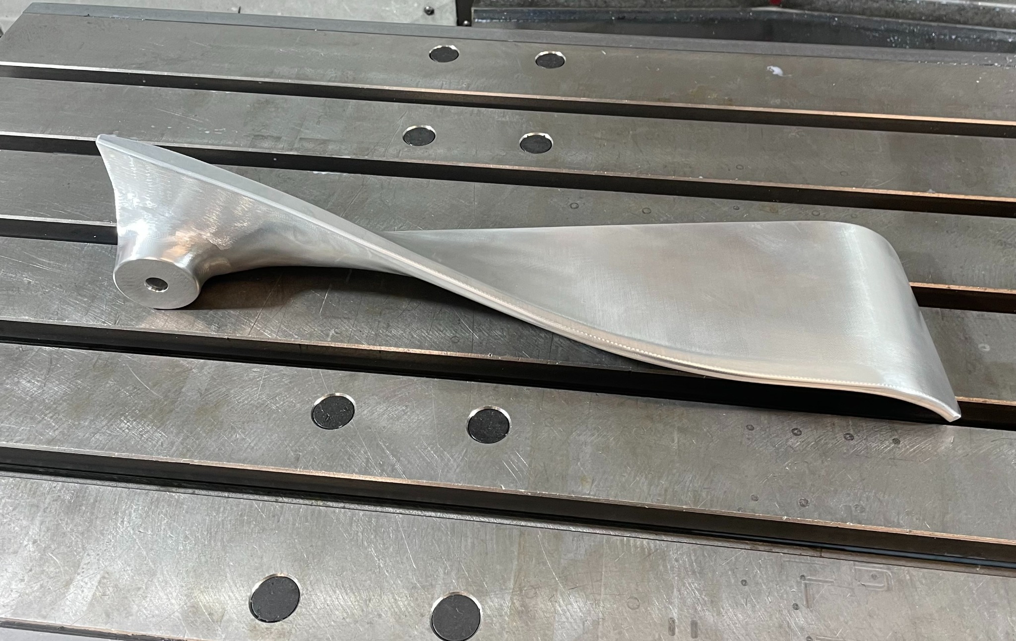 Metalowy, zakręcony element z otworem, leżący na stole frezarki. Widoczne ślady obróbki.