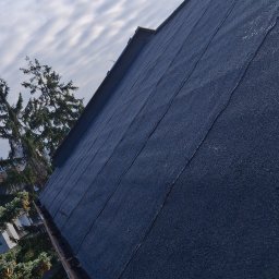 Adex - Dach pokryty papą, widok z dołu, rynna miedziana, fragment komina w tle, pochmurne niebo. Dach po wykonaniu prac dekarskich.