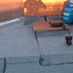 Adex - Szczegółowe ujęcie dachu pokrytego papą, z widocznym kominem, wentylacją i butlą gazową. Fragment starego dachu i nowa papa. Widoczne łączenia papy.