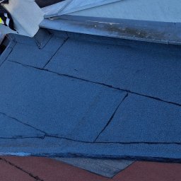 Adex - Widok z góry na fragment dachu pokrytego papą termozgrzewalną, z widocznym kominem z czerwonej cegły i niebieską butlą gazową. W tle zabudowa miejska.