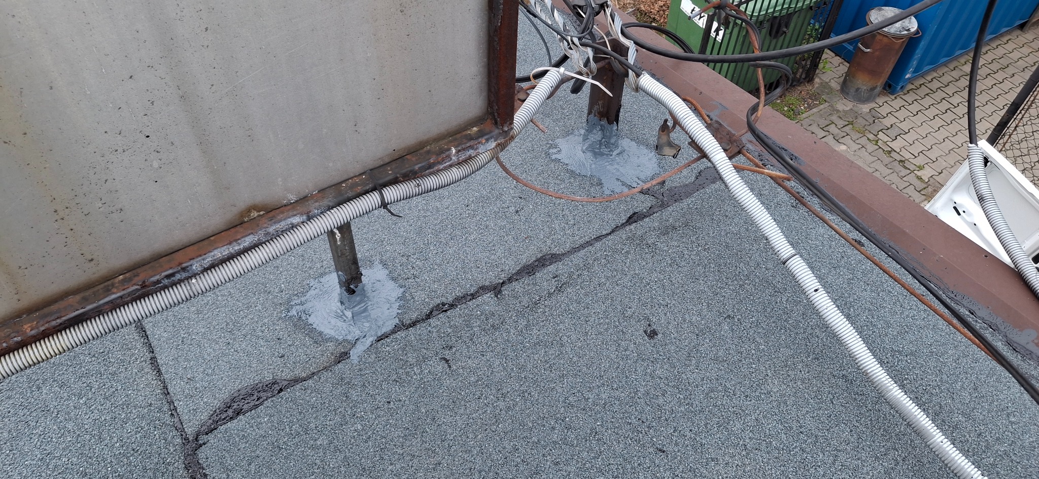 Uszczelnienie przejść instalacyjnych na dachu pokrytym papą, widoczne kable w peszlach i metalowa konstrukcja. Szare uszczelnienie wokół rur.