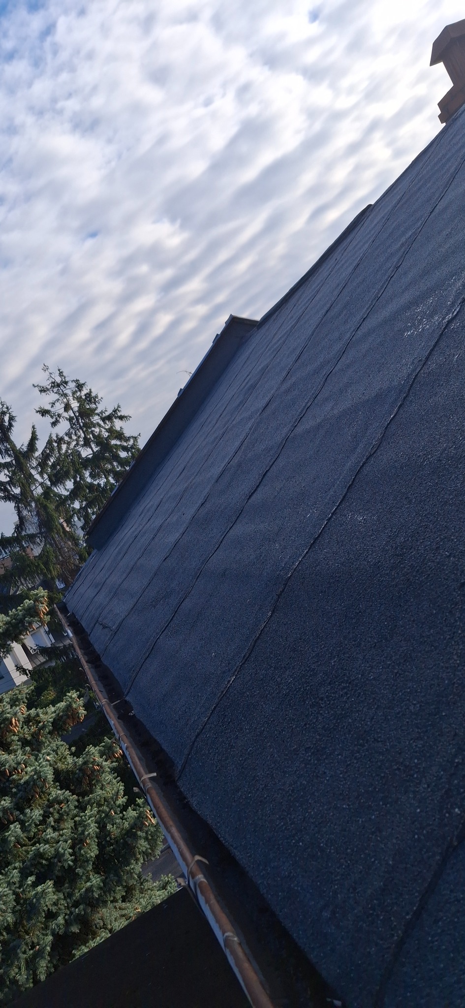 Dach pokryty papą, widok z dołu, rynna miedziana, fragment komina w tle, pochmurne niebo. Dach po wykonaniu prac dekarskich.