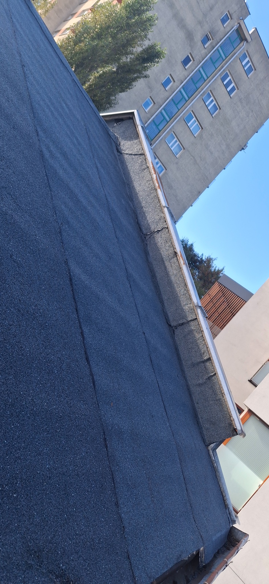 Ciemny, płaski dach pokryty papą, widoczny z boku, z fragmentem rynny i budynkami w tle. Układ połaci dachowej z wyraźnymi łączeniami. Widok na architekturę miejską i błękitne niebo.
