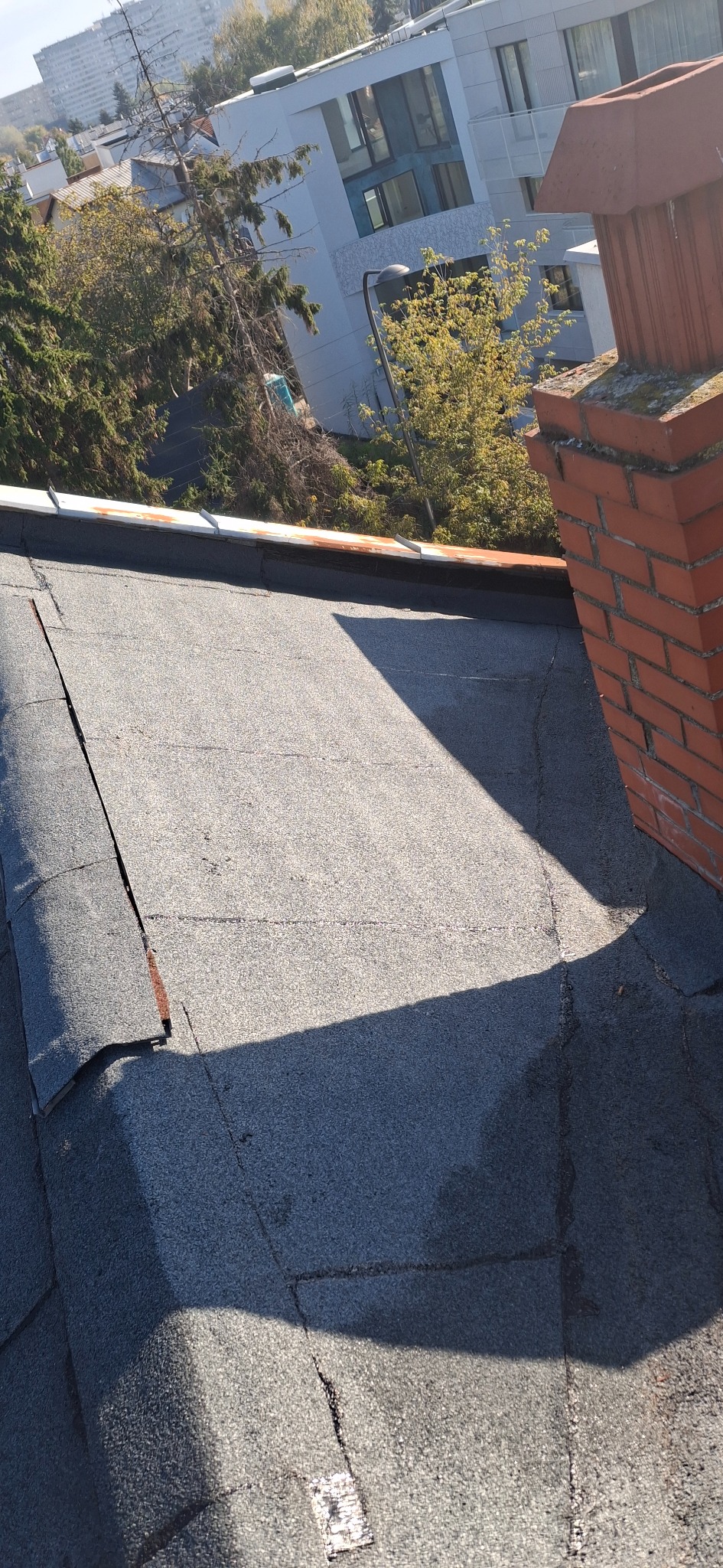 Uszkodzony, płaski dach z papy bitumicznej, widoczne pęknięcia i odspojenia. W tle komin z czerwonej cegły i budynki mieszkalne. Słoneczny dzień.