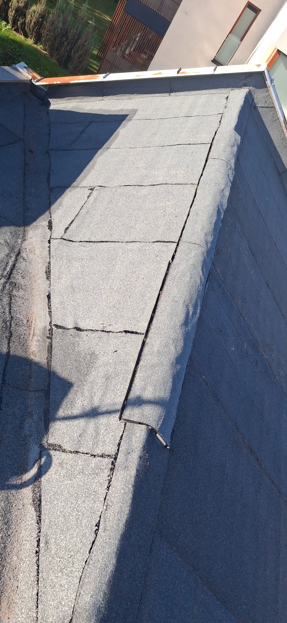 Uszkodzony dach pokryty papą z widocznymi pęknięciami i odklejonym fragmentem, ukazujący potrzebę naprawy. W tle fragment budynku i zieleni.