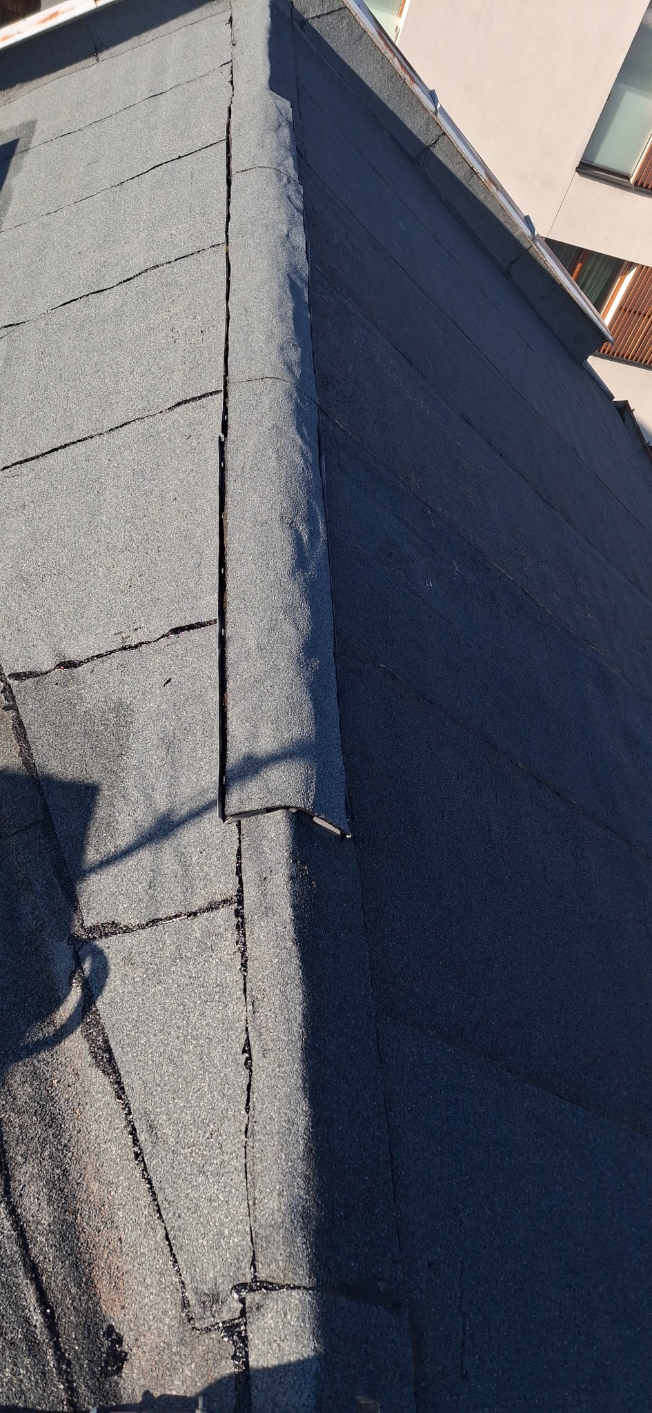 Uszkodzony dach pokryty papą, widoczne pęknięcia i nieszczelności. Fragment budynku w tle, cień na powierzchni dachu.