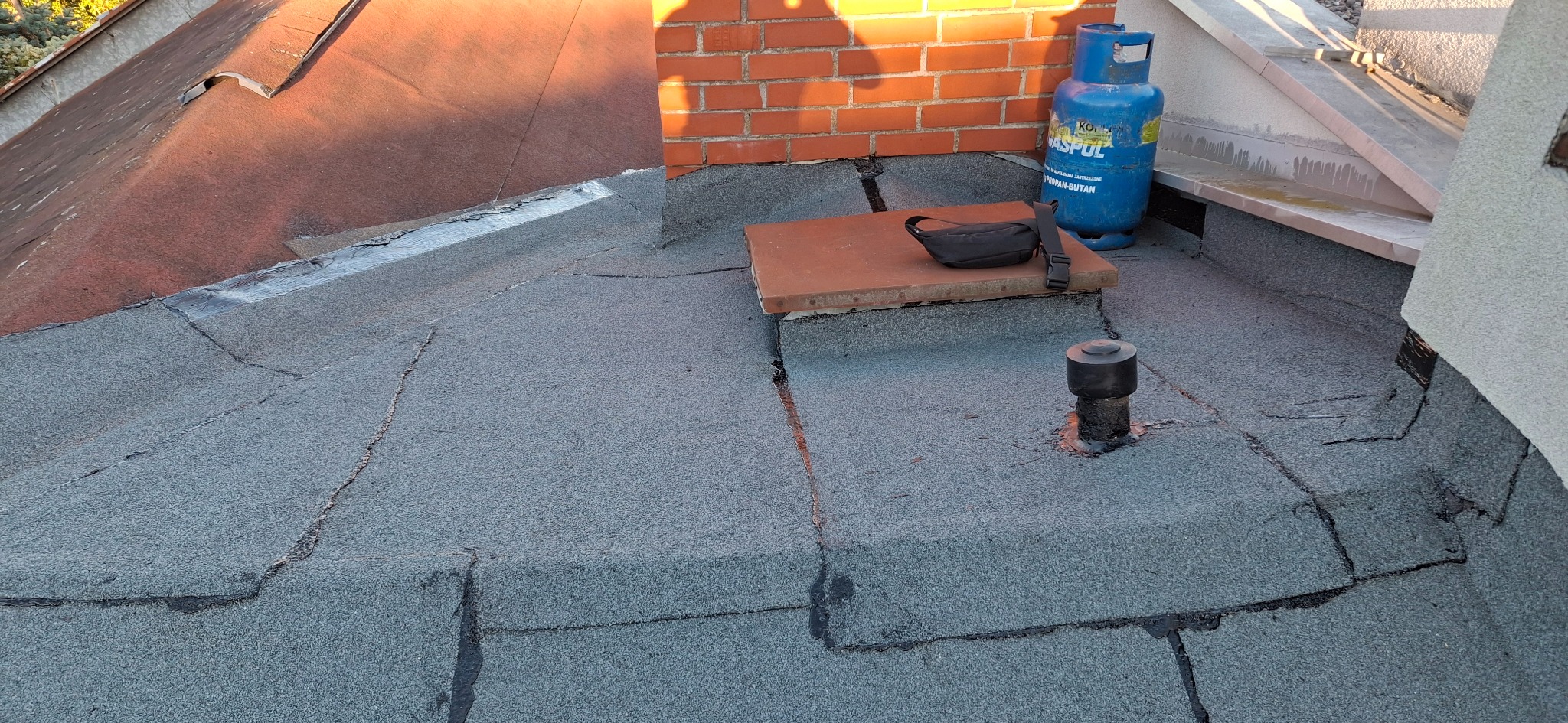 Szczegółowe ujęcie dachu pokrytego papą, z widocznym kominem, wentylacją i butlą gazową. Fragment starego dachu i nowa papa. Widoczne łączenia papy.