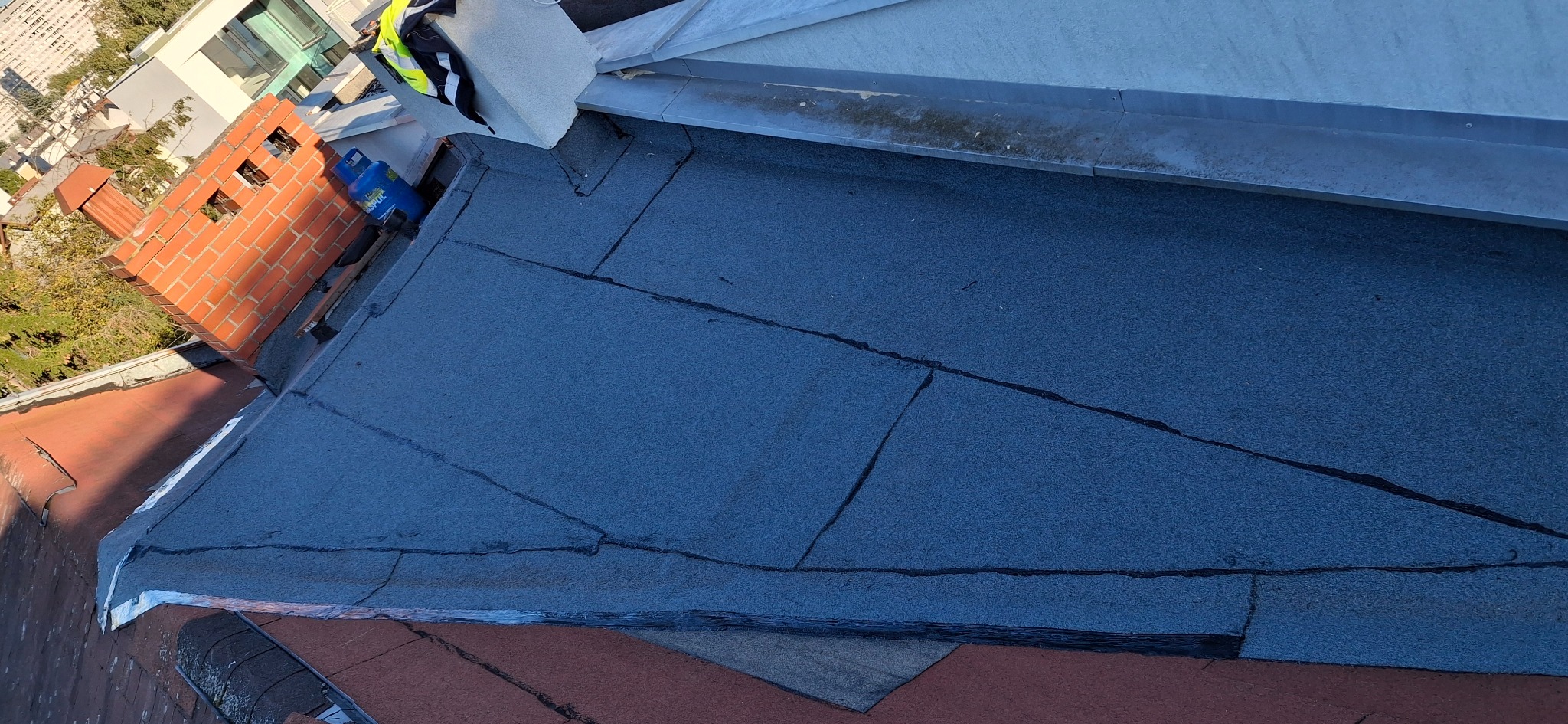 Widok z góry na fragment dachu pokrytego papą termozgrzewalną, z widocznym kominem z czerwonej cegły i niebieską butlą gazową. W tle zabudowa miejska.