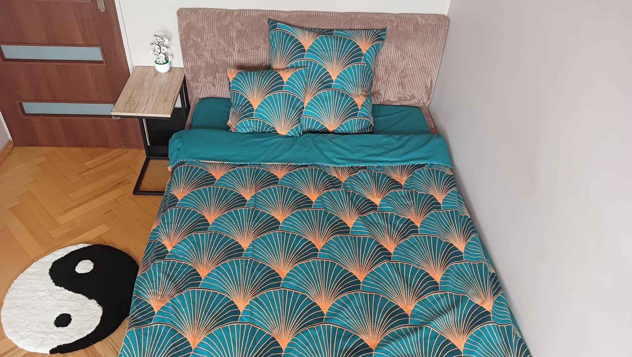 Komplet pościeli w turkusowo-złote wachlarze art deco na łóżku z beżowym zagłówkiem; obok stolik nocny z kwiatami i dywan Yin Yang.