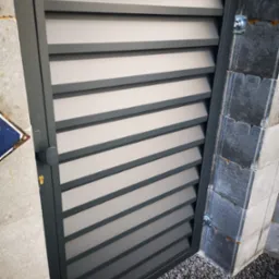 Nowoczesna, szara furtka metalowa z poziomymi panelami, zamontowana na murowanym słupku z kamienia łupanego, widoczna z bliska na tle szarego żwiru.