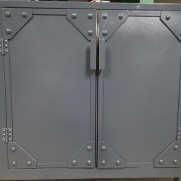 Mariusz Z. - Szara metalowa szafka dwudrzwiowa z nitowanymi narożnikami i prostymi uchwytami.