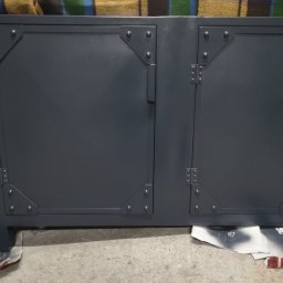 Mariusz Z. - Metalowa szafka z dwoma drzwiczkami, wzmocniona nitowanymi narożnikami, pomalowana na szaro, stoi na kartonach w warsztacie.