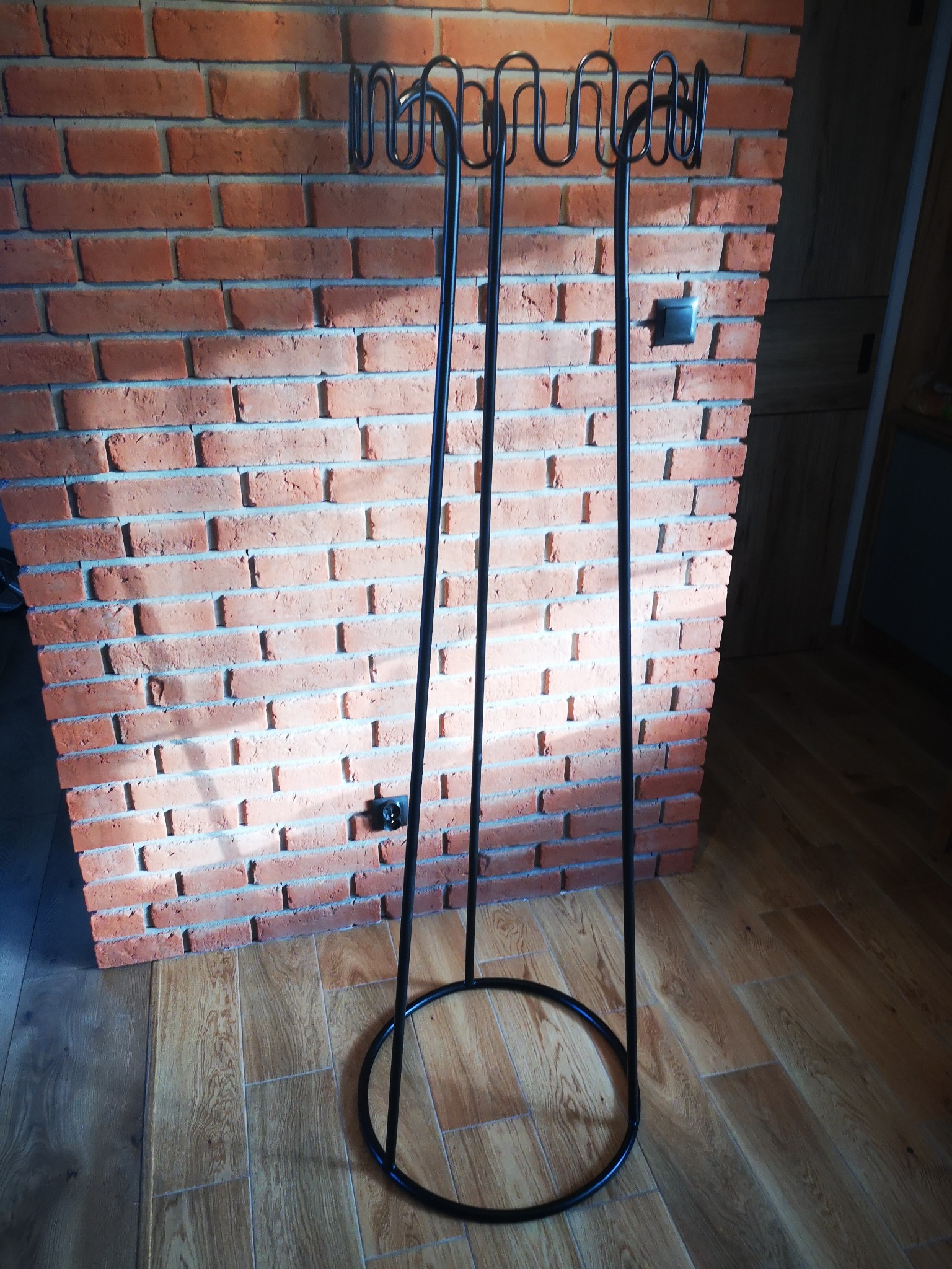 Metalowy stojak na drewno kominkowe, pomalowany na czarno, stoi przy ścianie z czerwonej cegły w nowoczesnym wnętrzu.