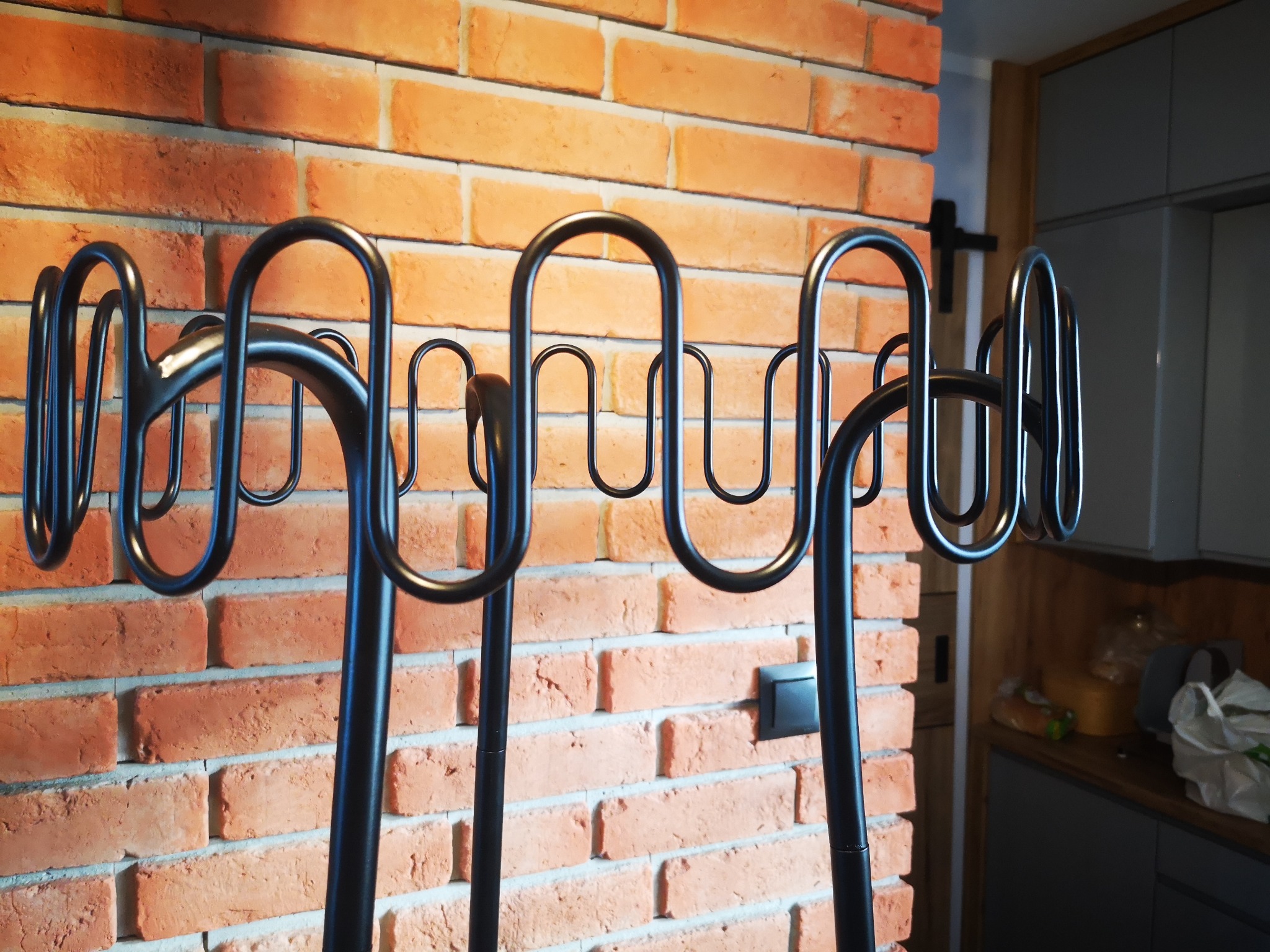 Czarny, metalowy stojak na ręczniki o falistym kształcie na tle ceglanej ściany w kuchni.