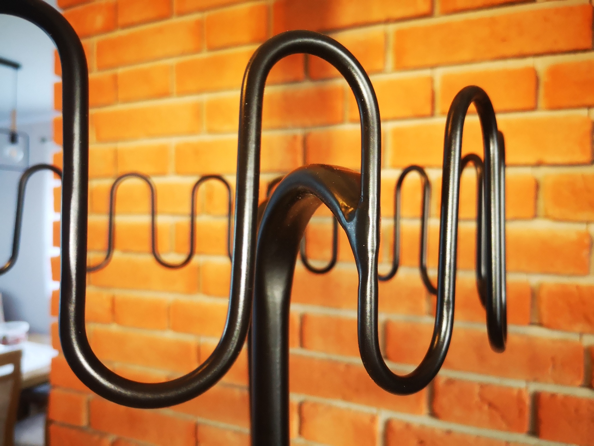 Czarny, metalowy wieszak na ubrania w stylu loft, z zakrzywionymi ramionami, na tle ściany z cegły klinkierowej w odcieniach pomarańczy i czerwieni. W tle widoczne elementy wyposażenia wnętrza.