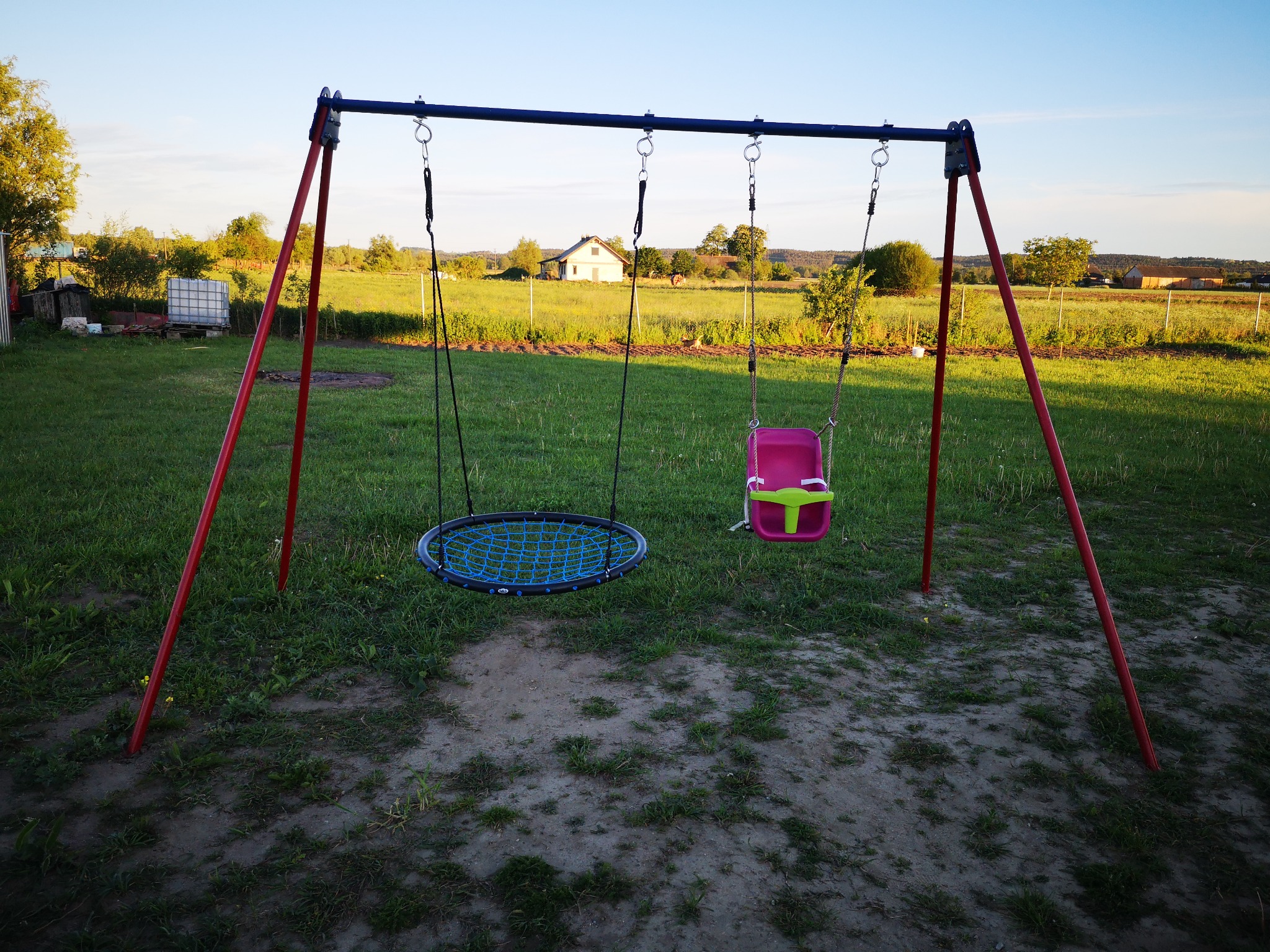 Metalowa konstrukcja huśtawki z okrągłym siedziskiem z siatki i plastikowym siedziskiem dla dziecka, ustawiona na trawniku przed domem na wsi.
