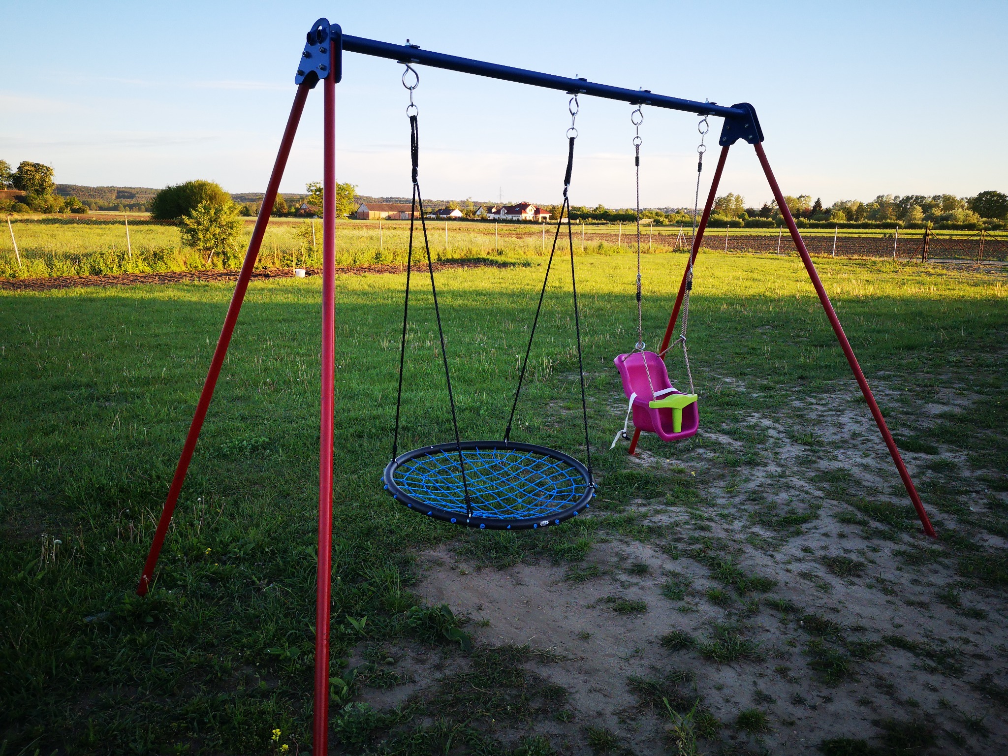 Metalowy stelaż huśtawki z dwoma siedziskami: okrągłym z niebieską siatką i różowym kubełkiem, na tle zielonego trawnika i pól uprawnych.