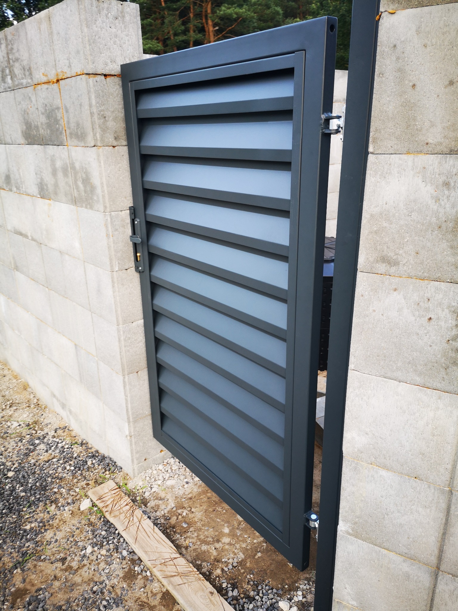 Nowoczesna, szara furtka z poziomymi lamelami, zamontowana na betonowym murze z bloczków, widoczny mechanizm zamka i zawiasy.