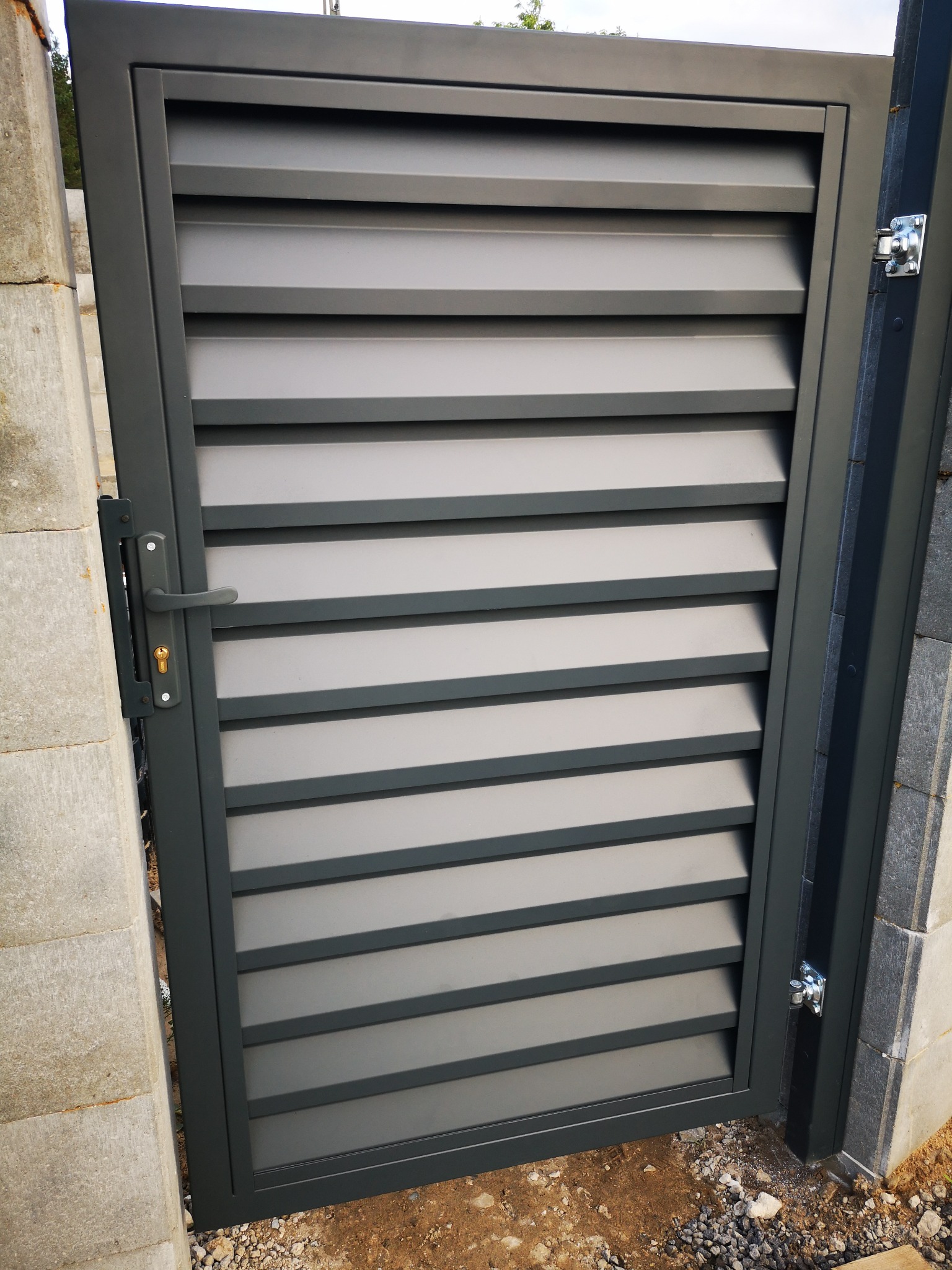 Nowoczesna, grafitowa furtka metalowa z poziomymi lamelami, zamontowana na betonowym słupku, widoczny zamek i zawiasy.