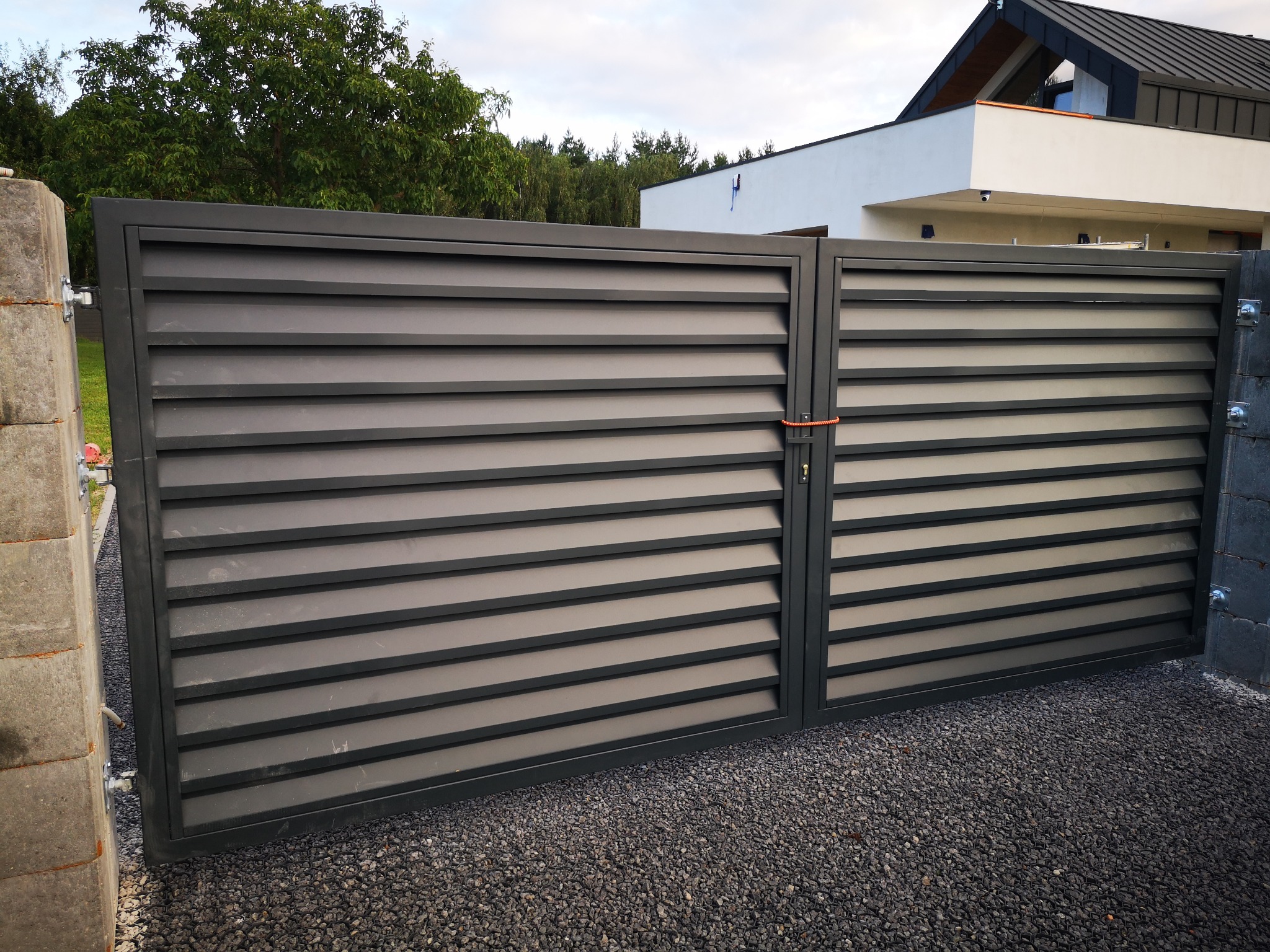 Nowoczesna, metalowa brama wjazdowa z poziomymi panelami, zabezpieczona pomarańczowym sznurkiem, na tle domu z białymi ścianami i szarym dachem, oraz podjazdu wysypanego szarym żwirem.