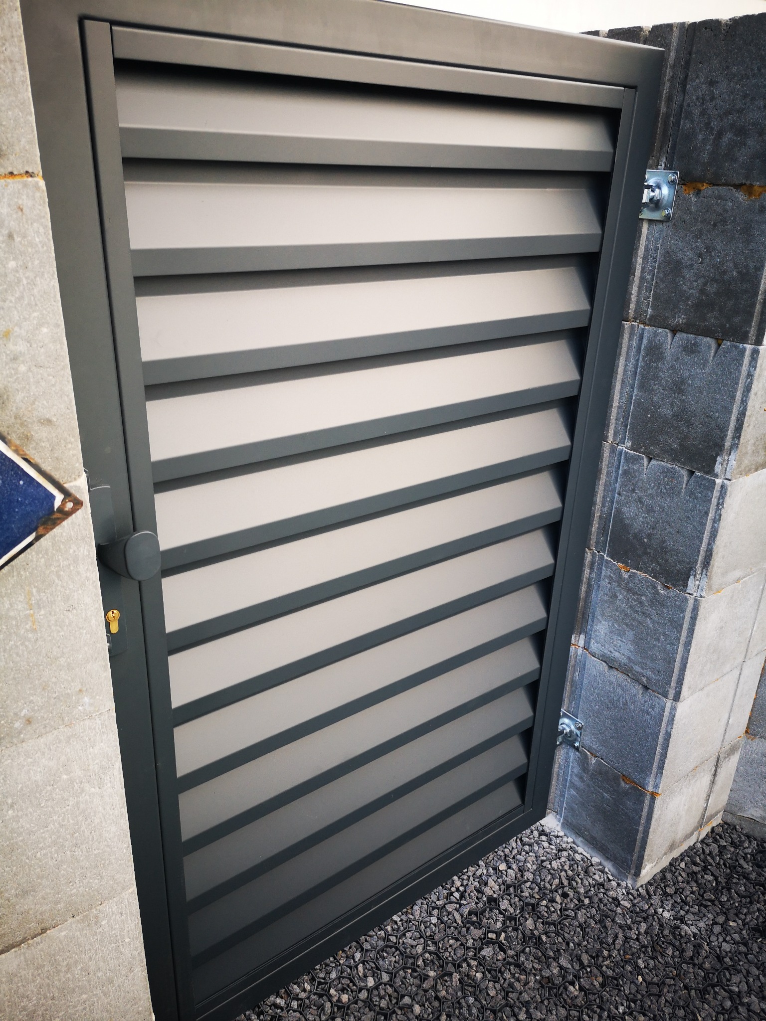 Nowoczesna, szara furtka metalowa z poziomymi panelami, zamontowana na murowanym słupku z kamienia łupanego, widoczna z bliska na tle szarego żwiru.