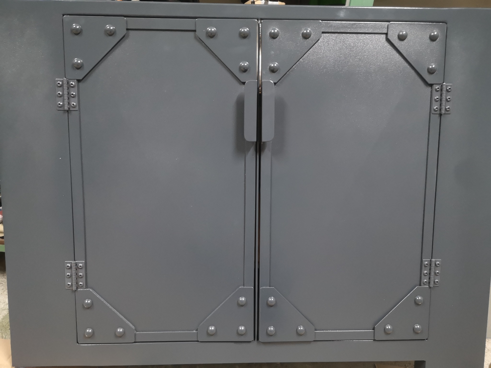 Szara metalowa szafka dwudrzwiowa z nitowanymi narożnikami i prostymi uchwytami.